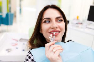 Woman in dental chair holding porcelain veneer color samples, smiling confidently, in a modern dental office setting.