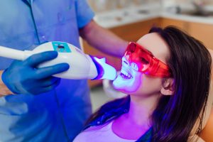 Patient undergoing professional teeth whitening with LED light activation, wearing protective eyewear and smiling during the procedure at Sweet Hart Dental.