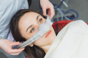 Woman receiving nitrous oxide sedation during dental treatment, wearing a mask for relaxation, with dental staff assisting, emphasizing anxiety relief techniques.